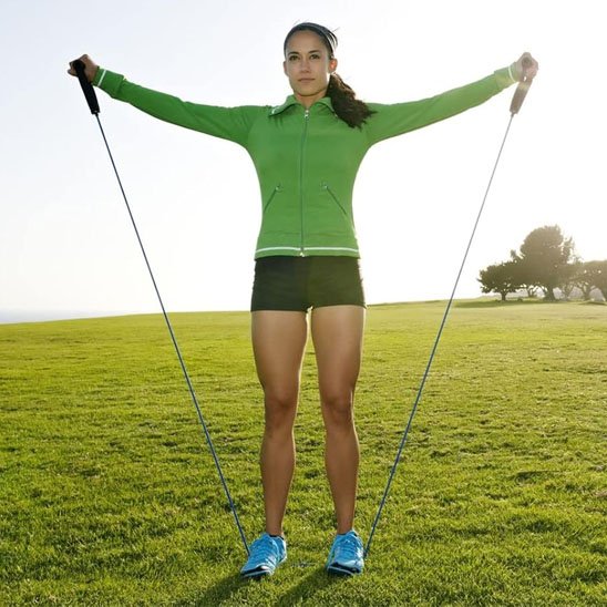 Femme étire élastique musculation en extérieur bras levés au soleil.